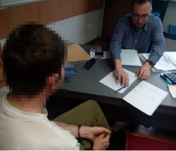 Fotografia kolorowa. Zatrzymany za włamanie sprawca przestępstwa siedzi przy biurku podczas przesłuchania przez funkcjonariusza kryminalnego.