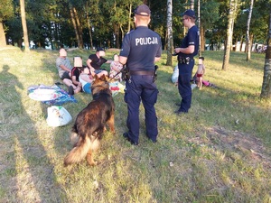 Dwóch odwróconych plecami policjantów z psem, w tle grupa ludzi