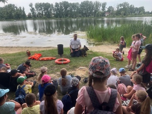 grupa dzieci słuchająca na siedząco policjanta nad jeziorem średzkim