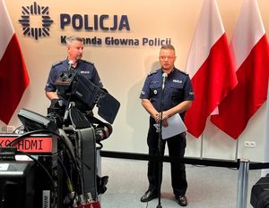 dwaj oficerowie policji w mundurach podczas briefingu na salce konferencyjnej stoją przed mikrofonami. Z przodu widać kamery. W tle na ścianie widnieje napis: Policja, Komenda Główna Policji , a z boku w stojakach stoją biało - czerwone flagi