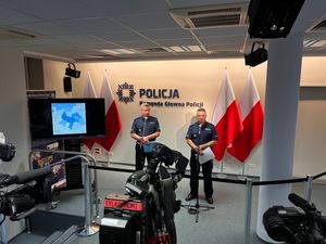 dwaj oficerowie policji w mundurach podczas briefingu na salce konferencyjnej stoją przed mikrofonami. Z przodu widać kamery. W tle na ścianie widnieje napis: Policja, Komenda Główna Policji , a z boku w stojakach stoją biało - czerwone flagi