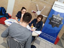 alt=&amp;quot;Fotografia kolorowa. Na pierwszym planie młody chłopak, który siedzi przy stoisku informacyjnym dotyczącym pracy w Policji. Po przeciwnej stronie siedzą przedstawicieli komendy w Środzie. W tle baner z logo jednostki.&amp;quot;