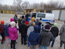 Fotografia kolorowa. Policjant z dziećmi przy radiowozie, pokazuje wyposażenie.