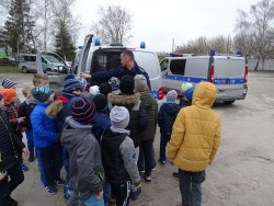 Fotografia kolorowa. Policjant pokazuje dzieciom w radiowozie miejsce do przewożenia zatrzymanych.